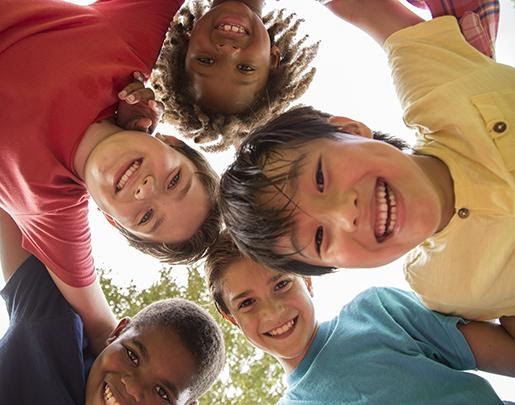 Small version of Image looking up towards a group of diverse kids standing arm in arm in a huddle, smiling down at camera for mobile